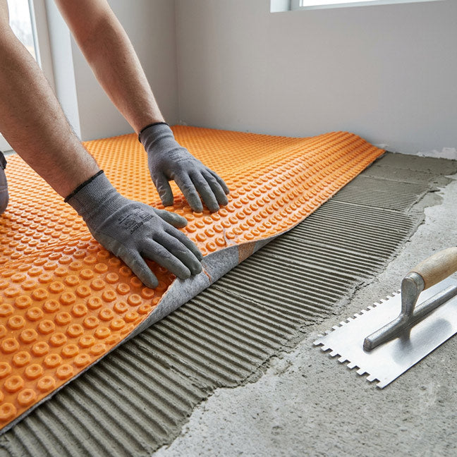 Step 1 — Gloved hands pressing DITRA-HEAT membrane onto a freshly comb-troweled bed of thinset