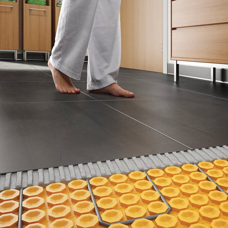 Step 3 — Bare feet on heated tile floor with cutaway revealing the DITRA-HEAT membrane and cable system underneath
